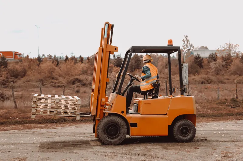 Heftruckchauffeur in oranje hesje rijdt op een buitenterrein met een heftruck naar een stapel pallets