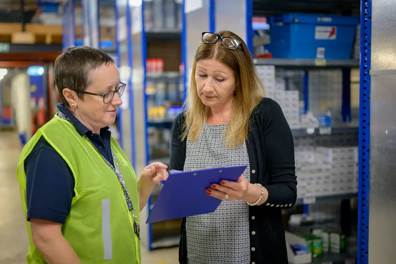 Leidinggevende in veiligheidshesje bespreekt met een collega een klembord in het magazijn