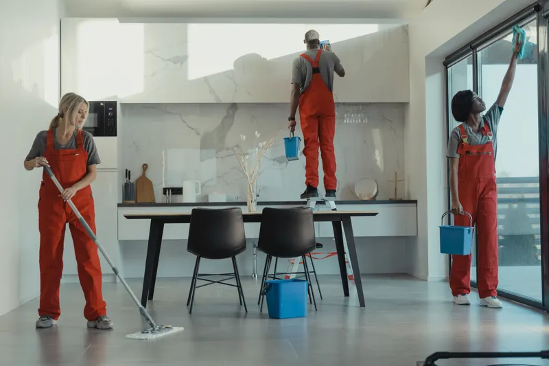 Drie schoonmakers in oranje overalls dweilen en lappen ramen in een lichte ruimte