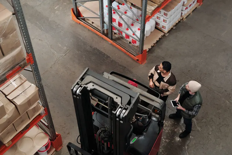 Teamleider met tablet overlegt met een medewerker naast een heftruck op de werkvloer van een magazijn