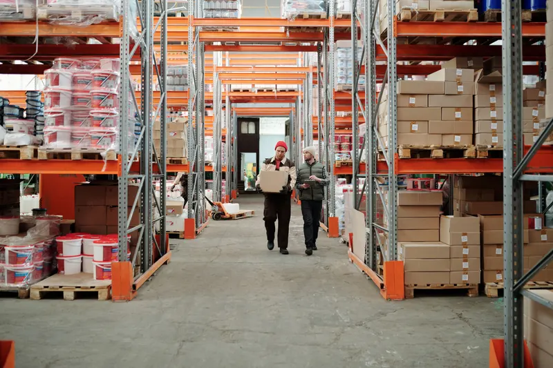 Twee collega's lopen met een doos door een gangpad tussen hoge stellingen in een magazijn
