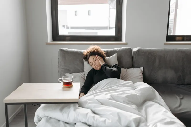 Zieke vrouw rust onder een dekbed op de bank, met een kop thee binnen handbereik